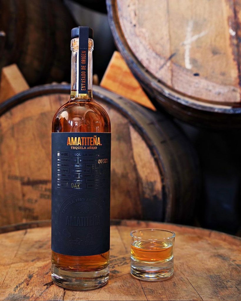 Amatiteña Añejo tequila bottle and tasting glass beside oak barrels